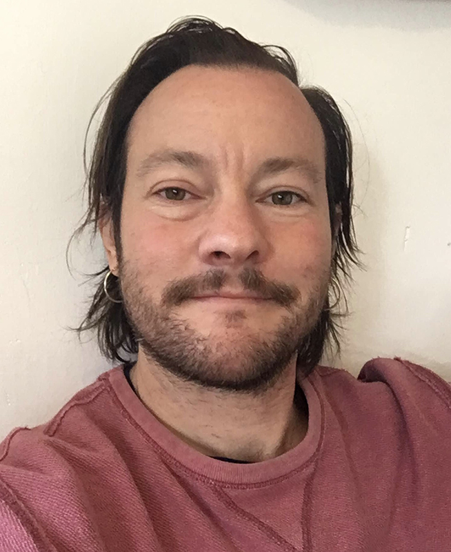 Christoph Hanssmann (Women and Gender Studies) A light-skinned person with a short brown beard and mustache smiles a closed mouth smile straight into the camera. They have short-to-mid length hair and are wearing a dusty red-colored t-shirt.