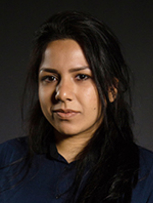 Maryam Khan (Engineering) A brown-skinned person with long black hair is in front of a grey backdrop. They are wearing a navy blue long sleeve collared shirt and have a serious look on their face.