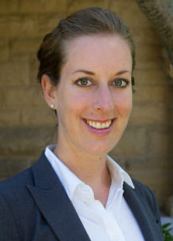 Meredith Reifschneider (Historical Archaeology, Anthropology) A smiling person with brown hair pulled back into a bun. They are in front of a yellow-brown brick wall. They are wearing diamond stud earrings and a navy blue blazer over a white collared shirt.