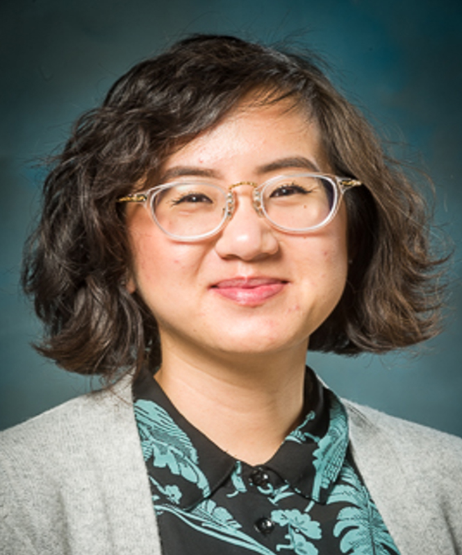 Michelle Tran (American Studies, Ethnic Studies, African American Studies & Sociology) A brown-skinned person with dark, wavy, chin-length hair smiles into the camera. They are wearing transparent glasses with gold accents. They have a black collared shirt with light greenish-blue leaves and vines under a gray cardigan. The backdrop they are in front of is the same hue as the leaves on their shirt.