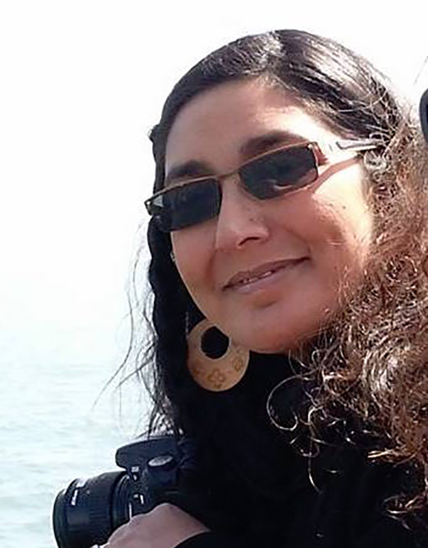 A brown-skinned person with shoulder-length, dark curly hair looks to their left into the camera and smiles. They are wearing a black jacket, brass-colored hoop earrings, and sunglasses. The sun is casting light on their right side, and there is a body of water behind them.