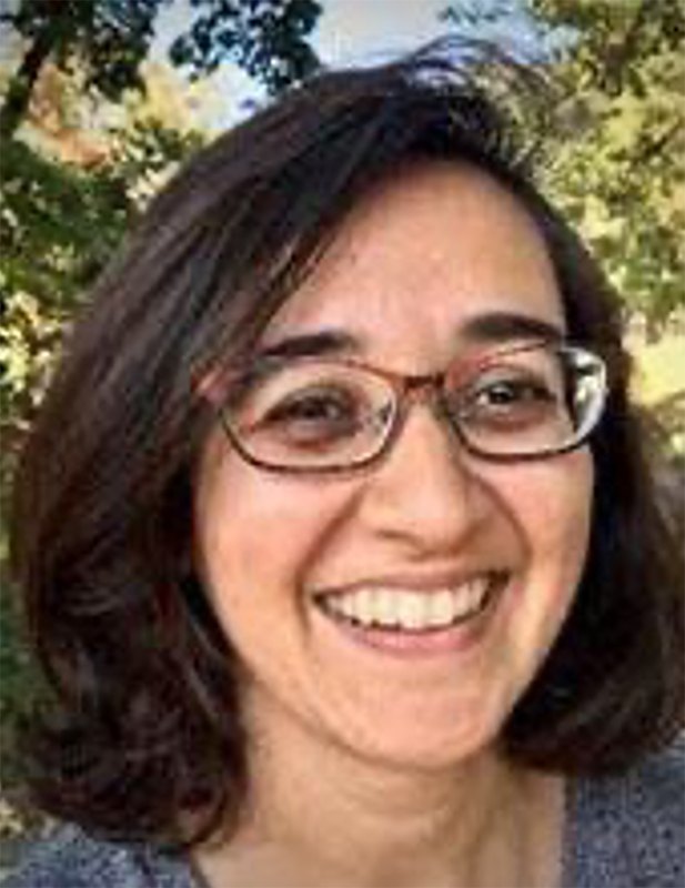 Supriya Misra (Public Health) A brown-skinned person with shoulder-length brown hair, curled at the bottom, is wearing glasses and smiling, looking to the camera's right. They are wearing a blue v neck top. There are deciduous trees with blue sky above them in the background. 