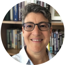 A smiling woman from the shoulders up, wearing glasses and a light blue button up. She has short salt-and-pepper hair and is in front of a bookshelf filled with books.