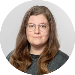 A woman wearing wire frame glasses and long, dark blonde hair from the shoulders up. Her shirt is dark green.